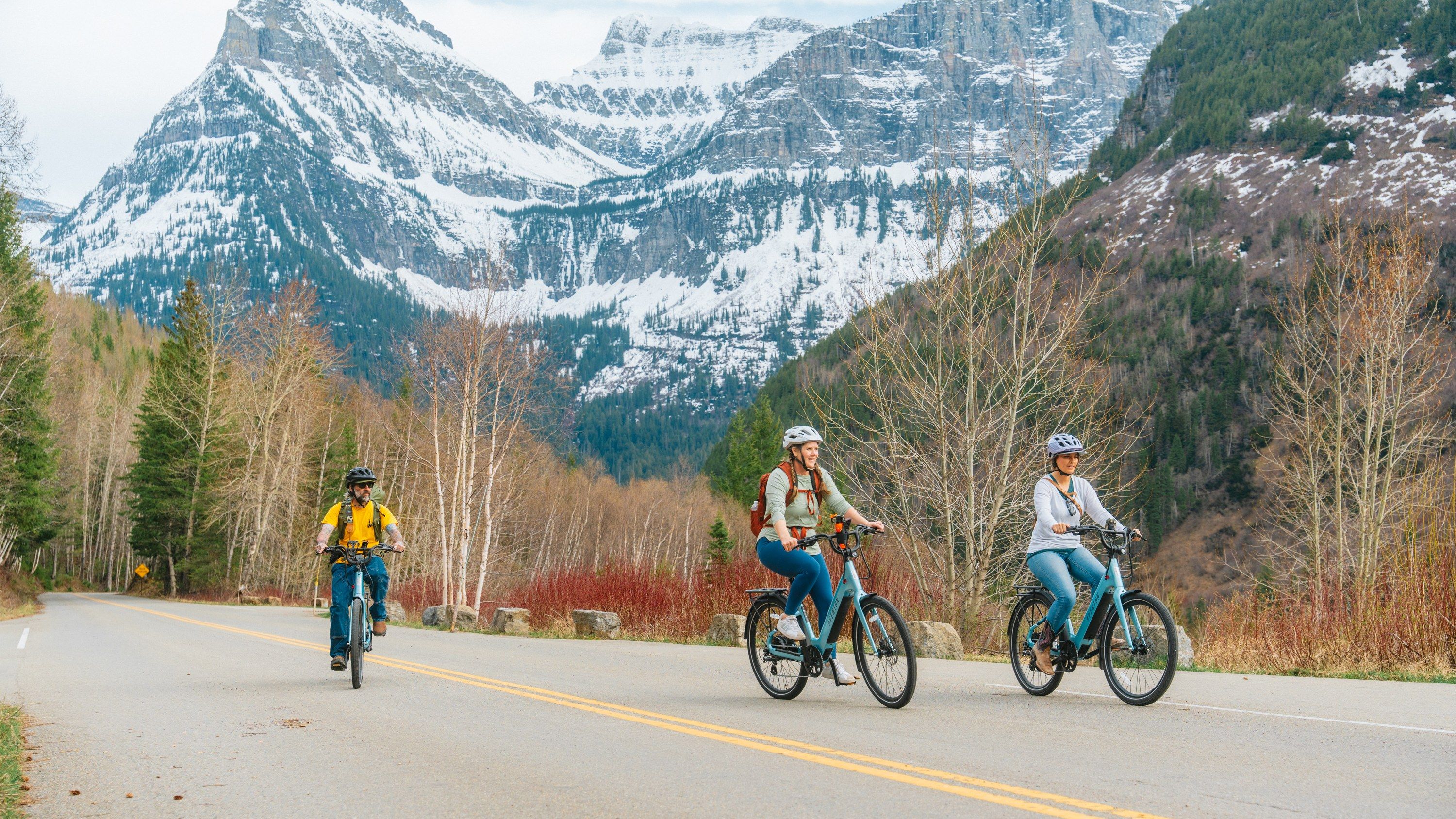 Three riders on e-bikes on Going-to-the-Sun Road below snowy peaks