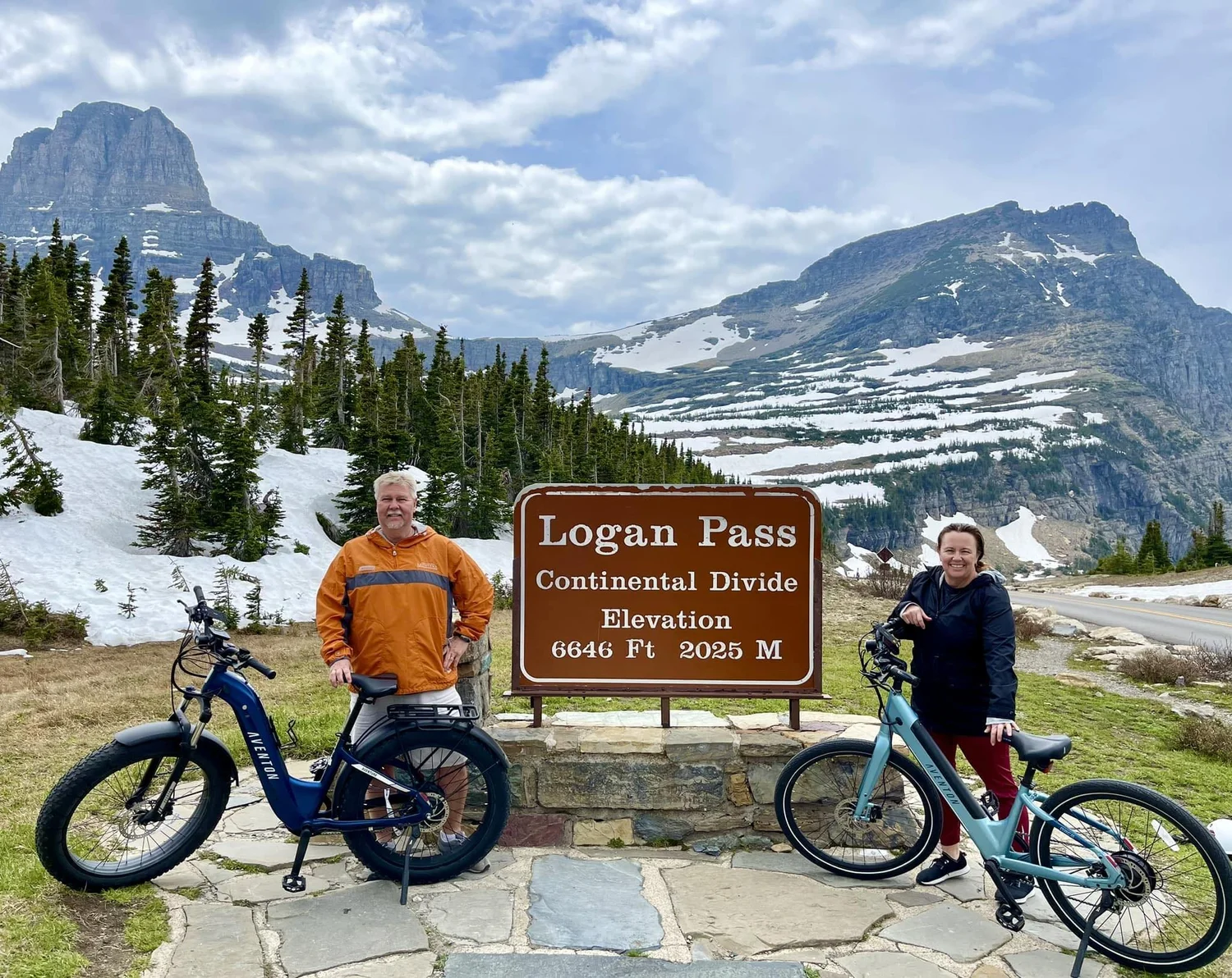 Two riders pose next to the Logan Pass sign at the top of Going-to-the-Sun Road after riding their e-bikes up the road